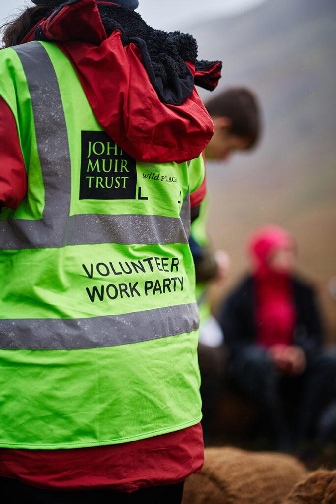 John Muir Trust volunteer wearing a hi-vis jacket with the John Muir Trust logo and volunteer work party on the back.